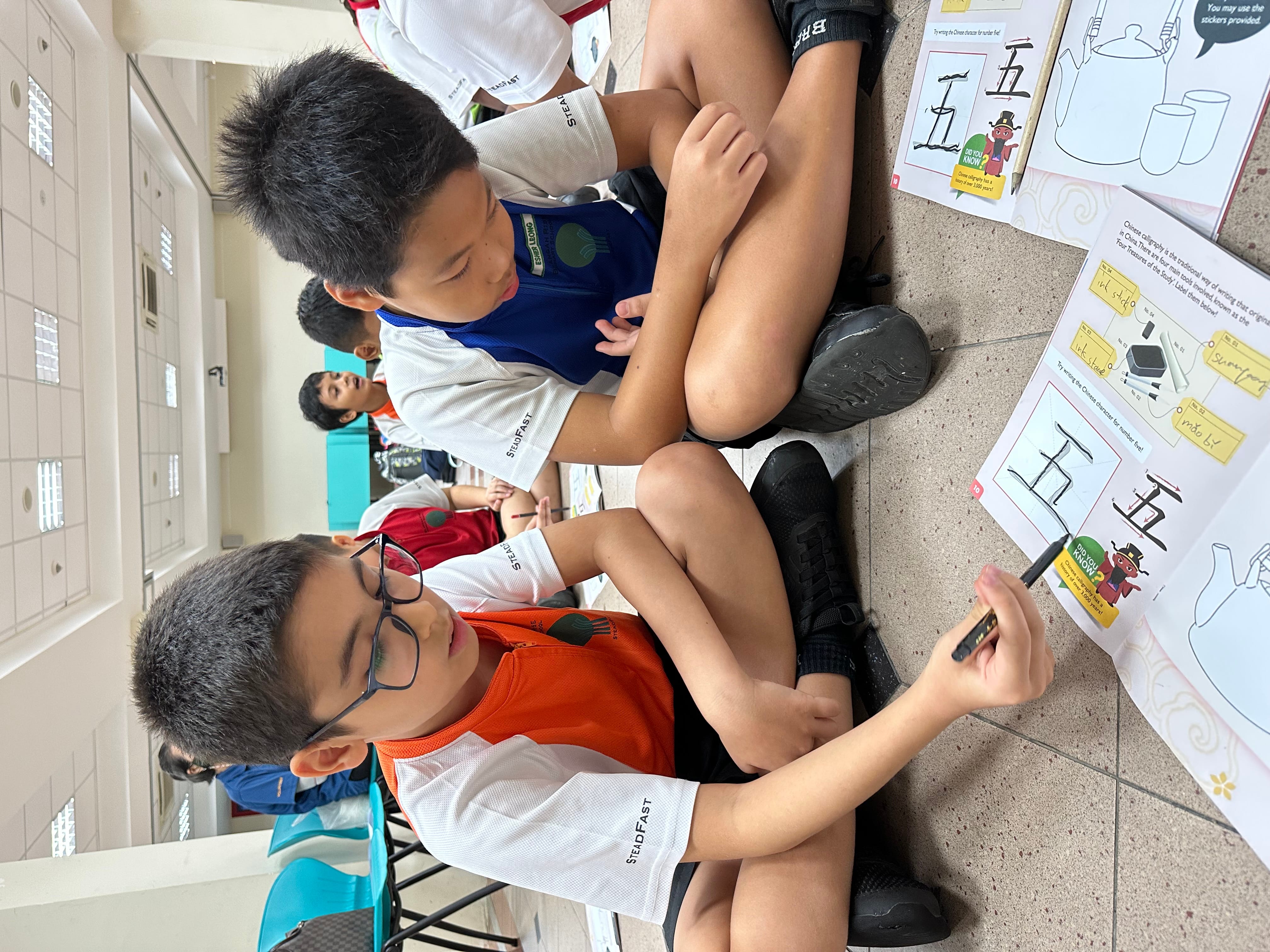 Two boys sit cross-legged on the floor doing a worksheet, one writing with a pencil.